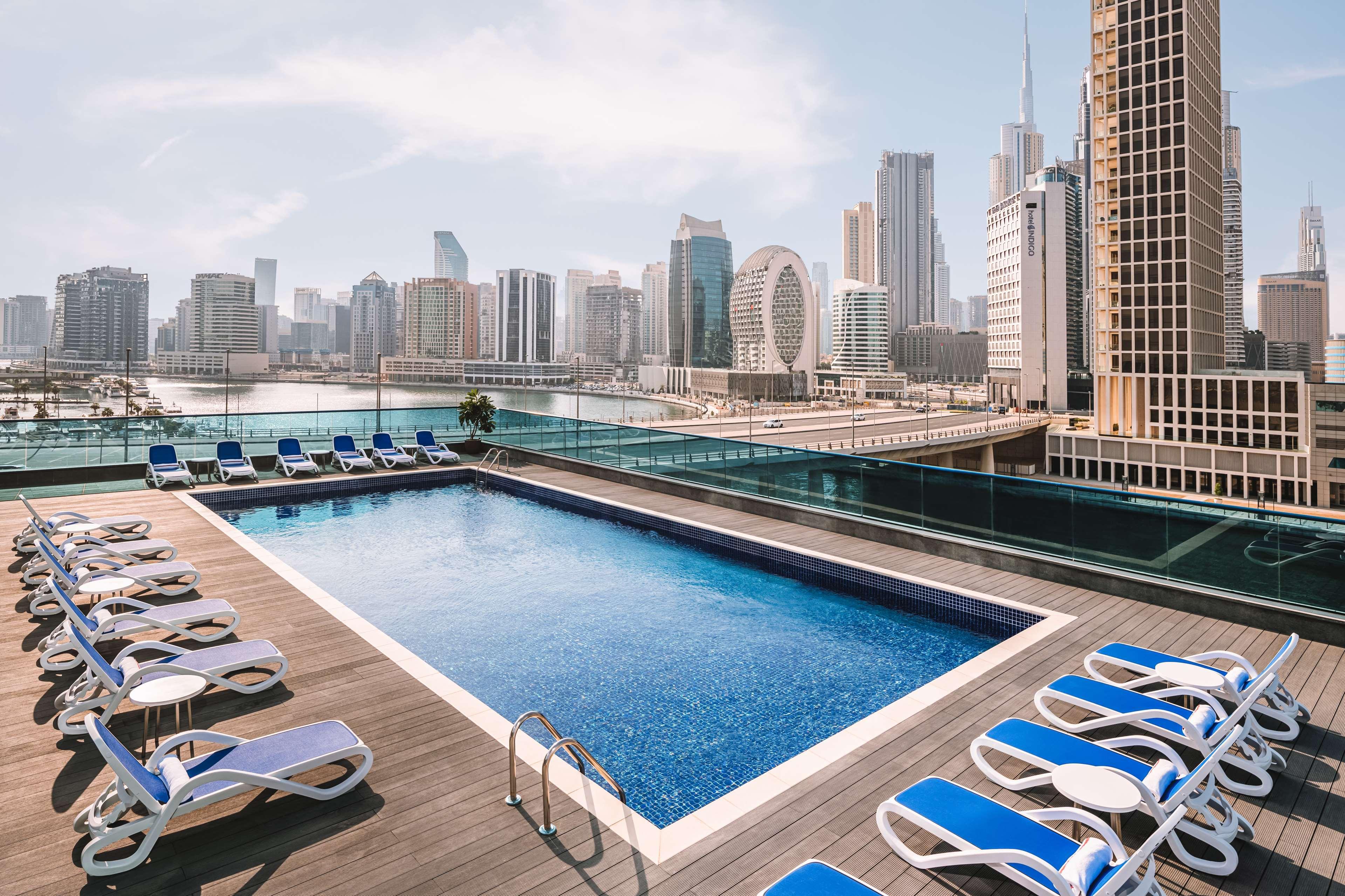 Radisson Blu Hotel, Dubai Canal View Exterior photo The image shows a modern rooftop pool area with a clear blue swimming pool surrounded by sun loungers. The loungers have blue cushions and are arranged neatly along the perimeter of the pool. In the background, there is a skyline of tall buildings, i