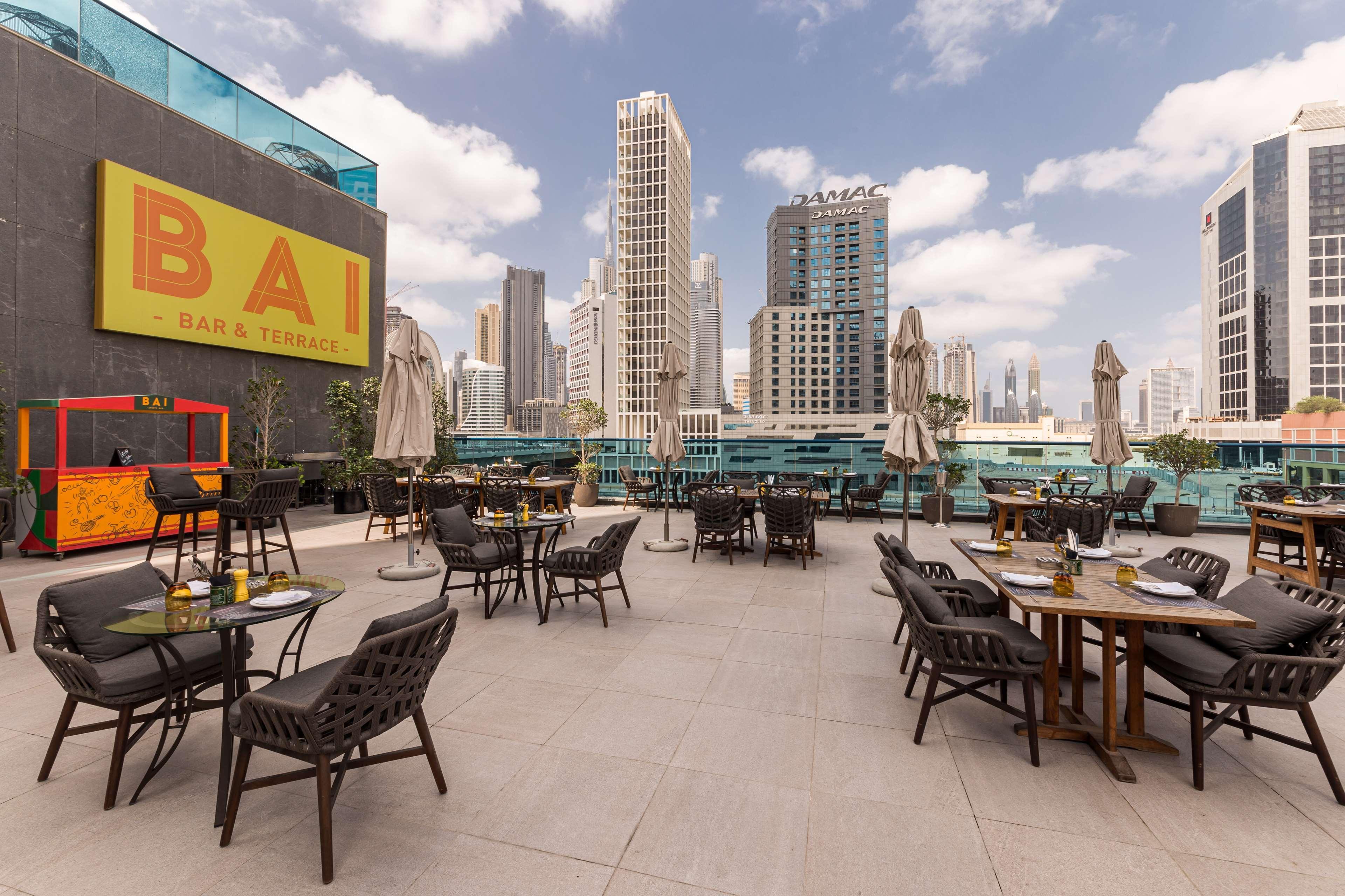 Radisson Blu Hotel, Dubai Canal View Exterior photo The photo shows a rooftop area of a bar or restaurant named "BAI," set against a backdrop of modern skyscrapers. There are several outdoor tables and chairs arranged on a paved surface, with some tables shaded by umbrellas. The space appears inviting